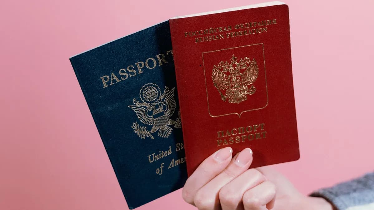 Hand holding a U.S. passport and a second passport against a pink background.