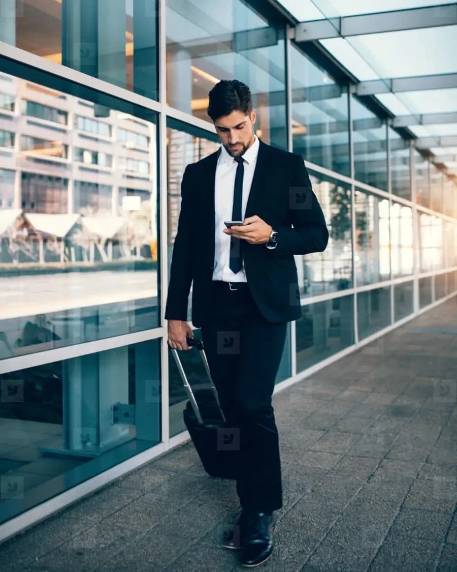 Business traveler with a rolling suitcase checking a phone outside a modern office building