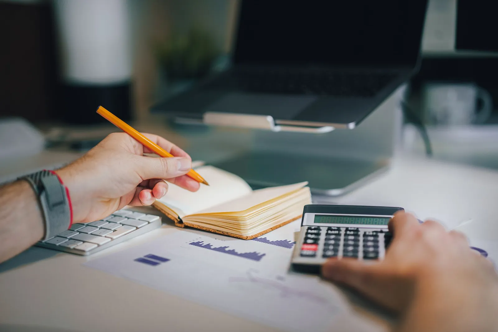 Hands taking notes beside a calculator and laptop while reviewing figures